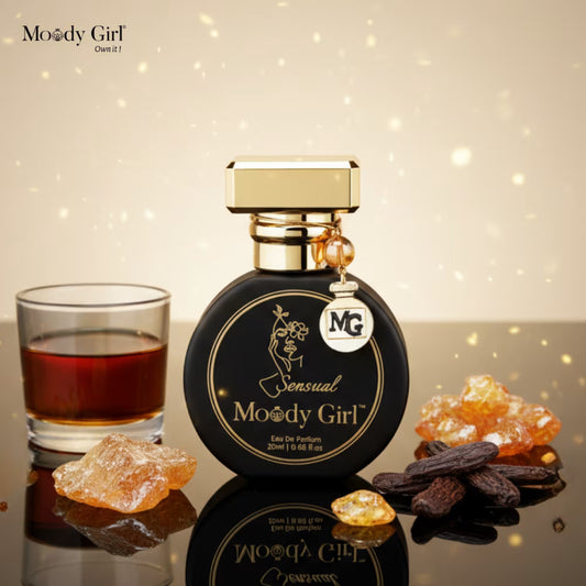 Perfume bottle labeled 'Moody Girl' with a glass of red liquid and amber stones on a reflective surface.