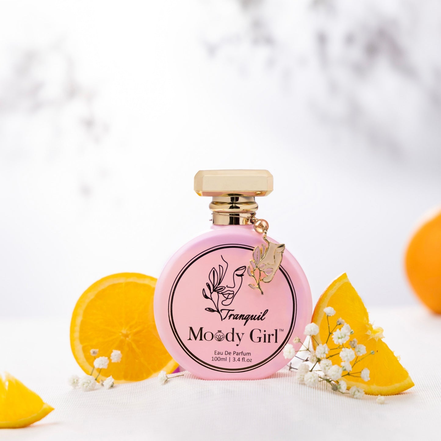 Tranquil 100ml Pink perfume bottle with gold cap labeled 'Moody Girl' on a white background with orange slices and flowers.