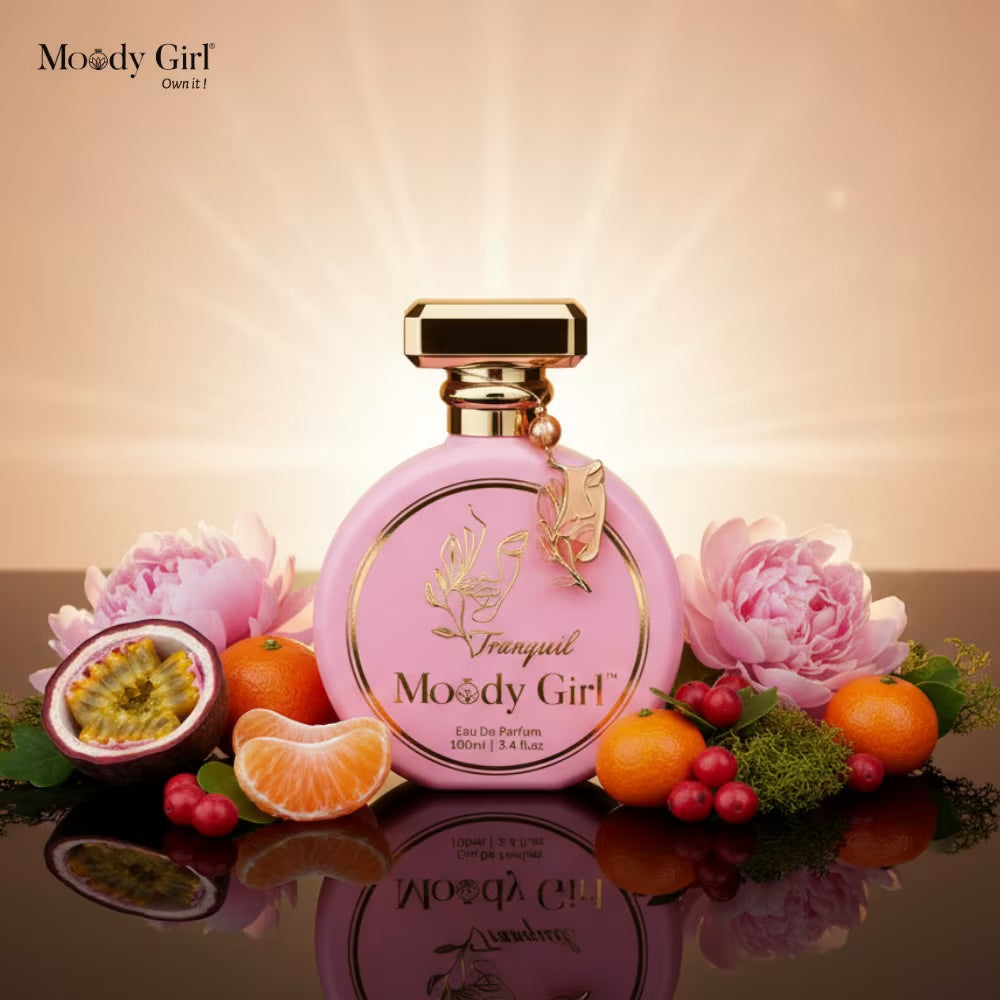 Pink perfume bottle with gold accents on a reflective surface with fruits and flowers, featuring 'Moody Girl' branding.