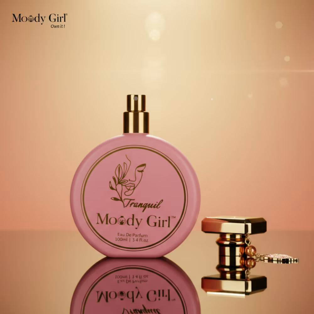 Pink perfume bottle with gold cap on a reflective surface against a warm gradient background, featuring 'Moody Girl' branding.