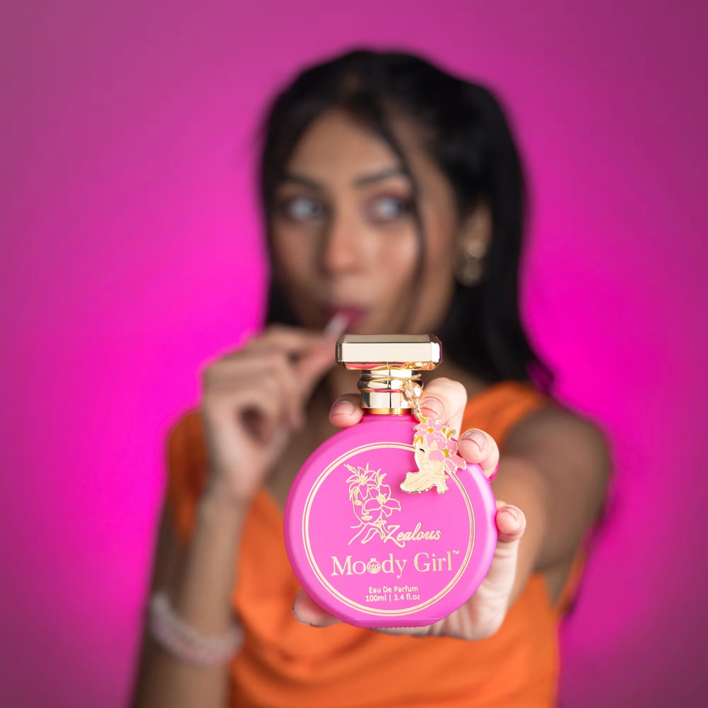 Person holding a pink 'Moody Girl' perfume bottle against a pink background