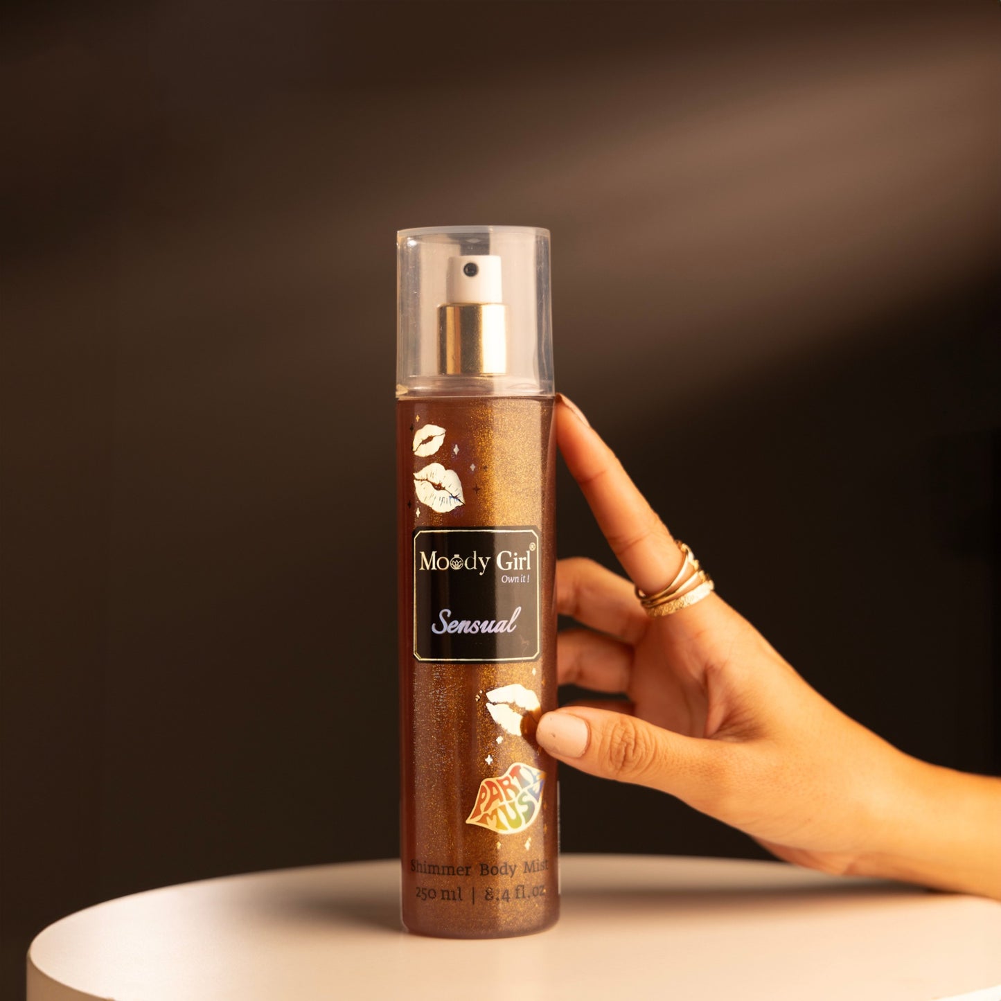 Hand holding a bottle of 'Moody Girl Sensual' body spray against a dark background