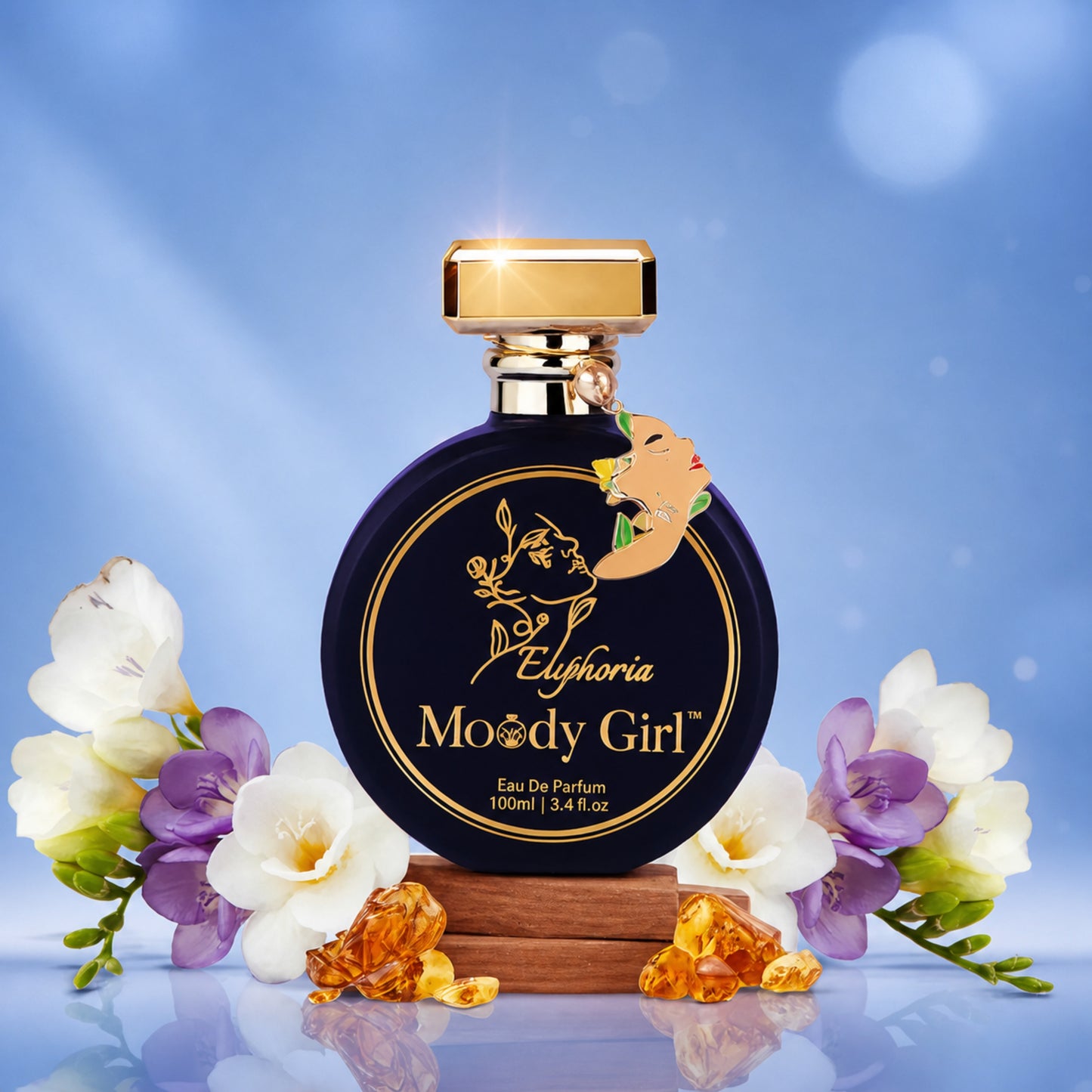 A round dark blue perfume bottle with a gold cap and gold lettering reading 'Euphoria Moody Girl' stands on a small wooden base, surrounded by white and purple flowers and amber crystal-like pieces against a soft blue background.