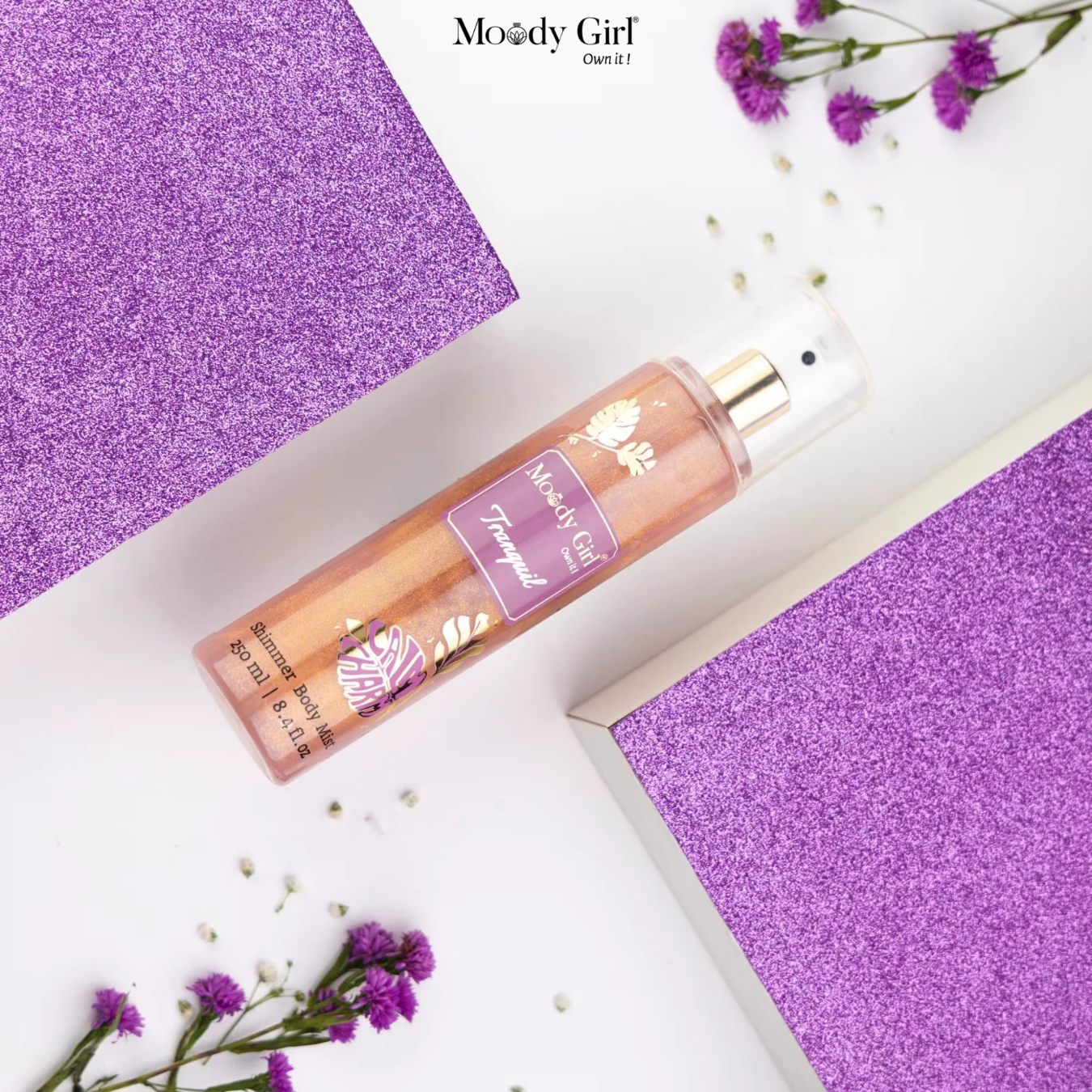 Moody Girl perfume bottle on a purple textured surface with flowers.