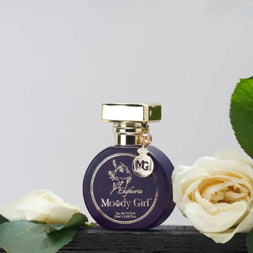 Perfume bottle labeled 'Moody Girl' with gold cap and decorative elements, surrounded by white roses on a light gray background.