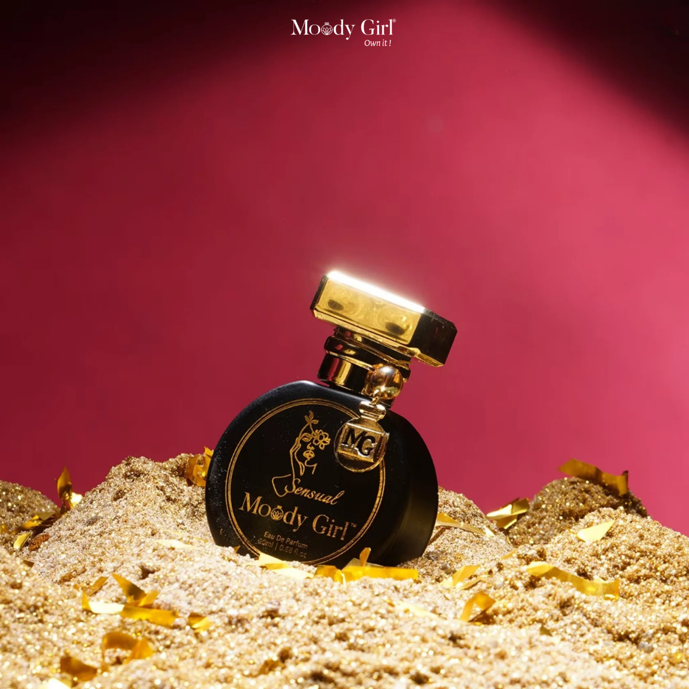 Perfume bottle labeled 'Moody Girl' on a sand-like surface with a red background