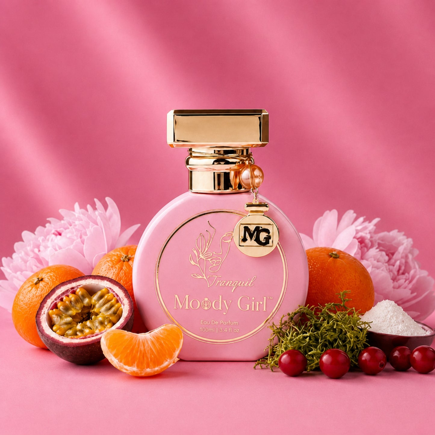 A pink perfume bottle with gold detailing and a hanging MG charm, styled on a pink background with oranges, a halved passion fruit, flowers, herbs, and red berries around it. The bottle label reads 'Vagual Moody Girl' and 'Los Angeles, CA'.