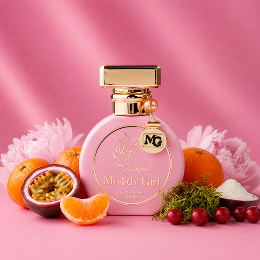 A pink perfume bottle with gold detailing and a gold cap, labeled 'Vanguished Moody Girl Eau de Parfum,' styled on a pink background with citrus fruits, flowers, and berries around it.
