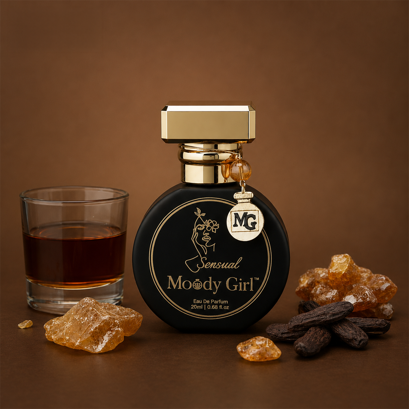 A bottle of Sensual Moody Girl Eau De Parfum with a gold cap and a hanging NG charm, styled on a brown background with a glass of amber liquid and amber crystals.