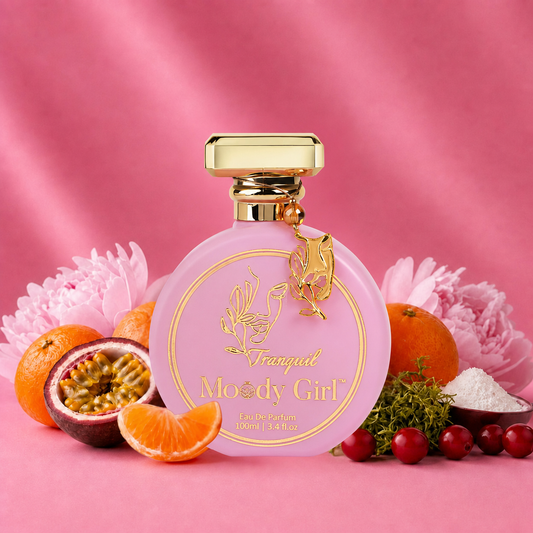 A pink perfume bottle with a gold cap and gold label reading 'Tranquil Moody Girl' is centered on a pink fabric backdrop, surrounded by citrus fruits, flowers, and berries as styled props.