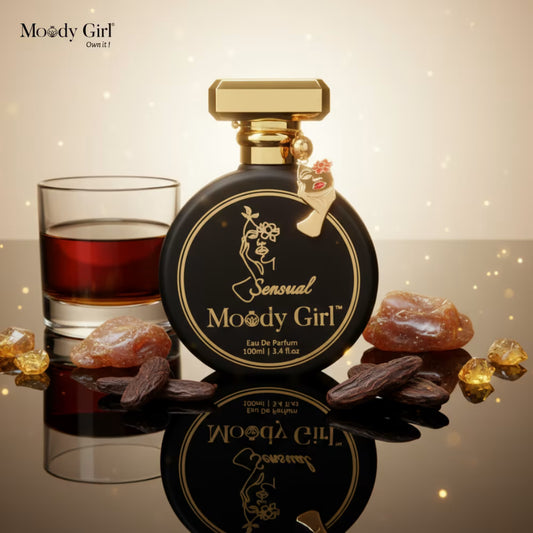 Perfume bottle labeled 'Sensual Moody Girl' with a glass of red rum and stones on a reflective surface.