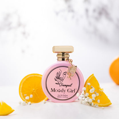 Tranquil 100ml Pink perfume bottle with gold cap labeled 'Moody Girl' on a white background with orange slices and flowers.