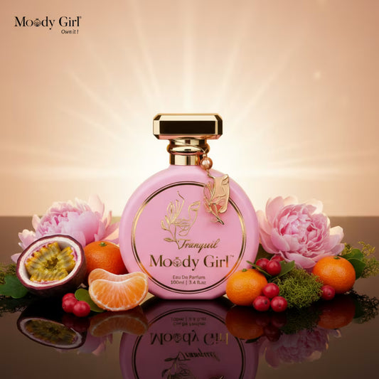 Pink perfume bottle with gold accents on a reflective surface with fruits and flowers, featuring 'Moody Girl' branding.