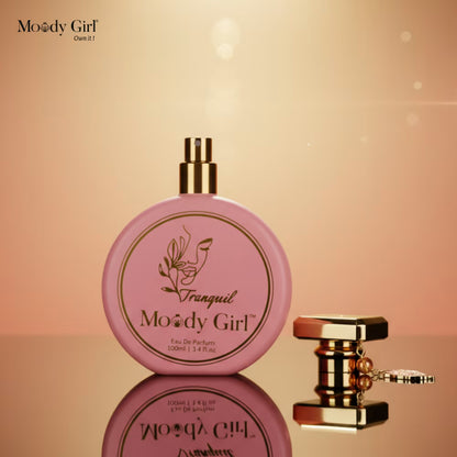 Pink perfume bottle with gold cap on a reflective surface against a warm gradient background, featuring 'Moody Girl' branding.