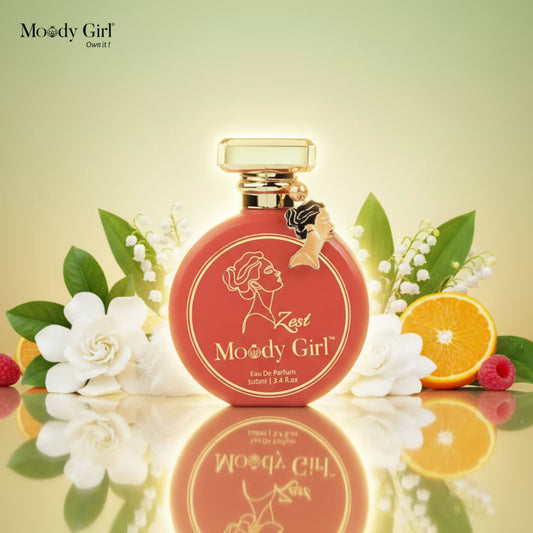 Red perfume bottle with gold cap labeled 'Moody Girl' surrounded by flowers and fruits on a reflective surface.