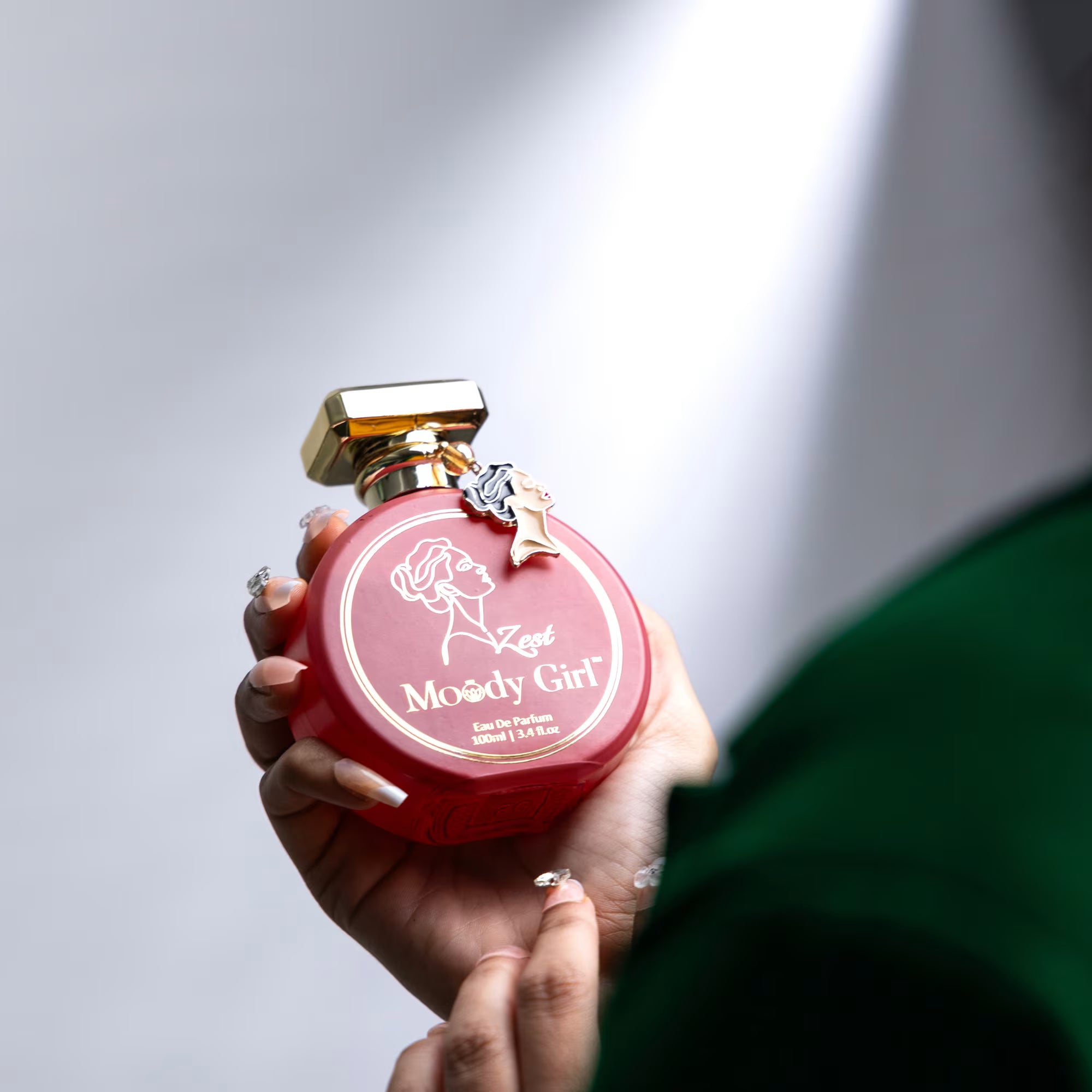 Hand holding a red 'Moody Girl' perfume bottle with gold cap against a neutral background