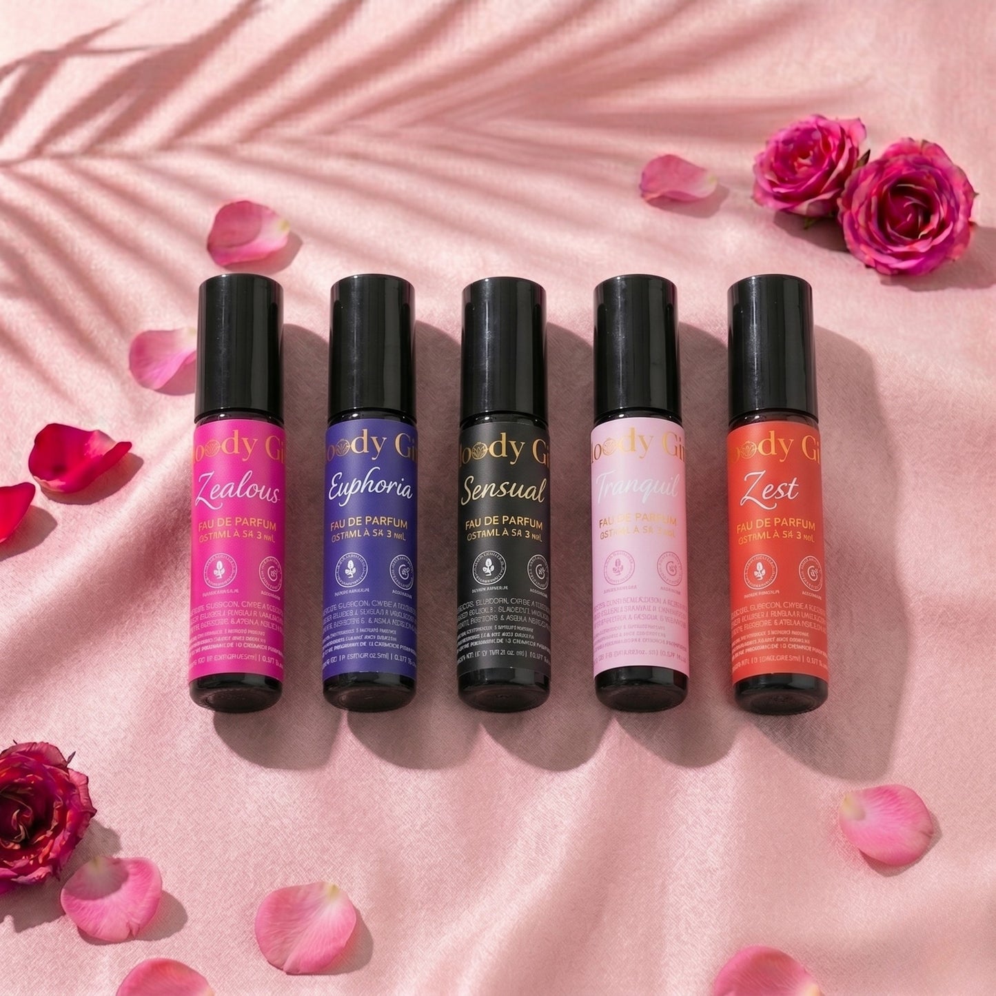 Five small perfume bottles with colorful labels on a pink fabric background with flowers and petals.