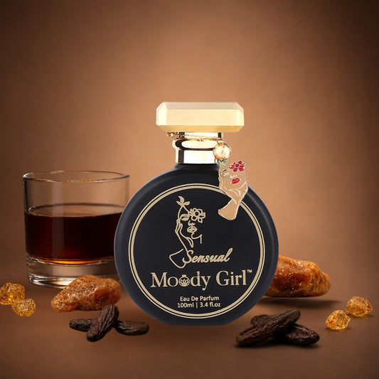 A bottle of Sensual Moody Girl Eau De Parfum is centered on a warm brown background, styled with a glass of dark whiskey, amber resin pieces, and dark spices. The bottle has a round black label with gold lettering and a cream-colored cap.