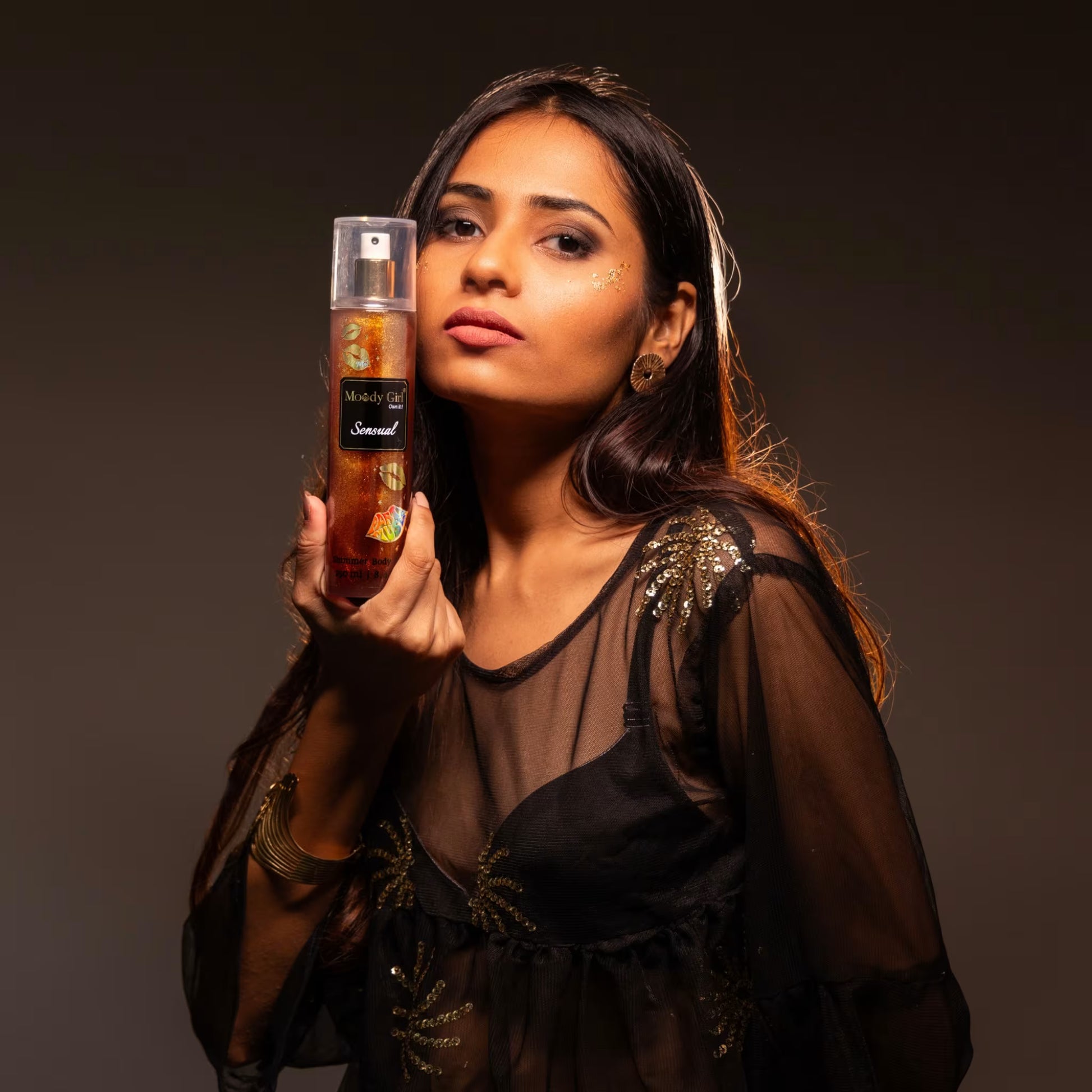 A woman holding a bottle of Moody Girl Sensual Shimmer Body Mist against a dark background