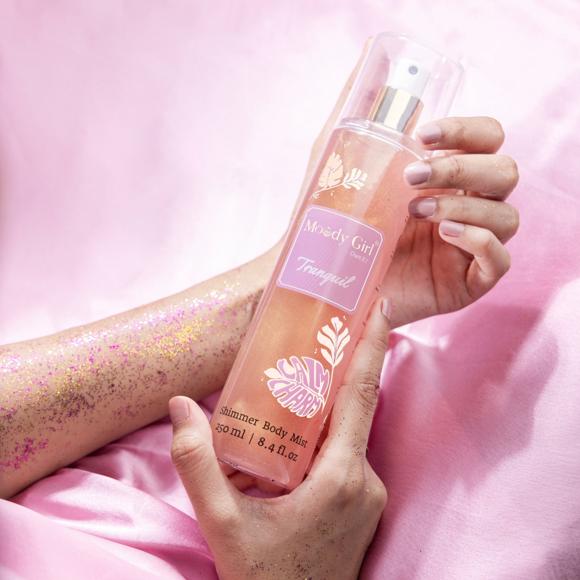 Hand holding a 'Moody Girl' shimmer body mist against a pink background