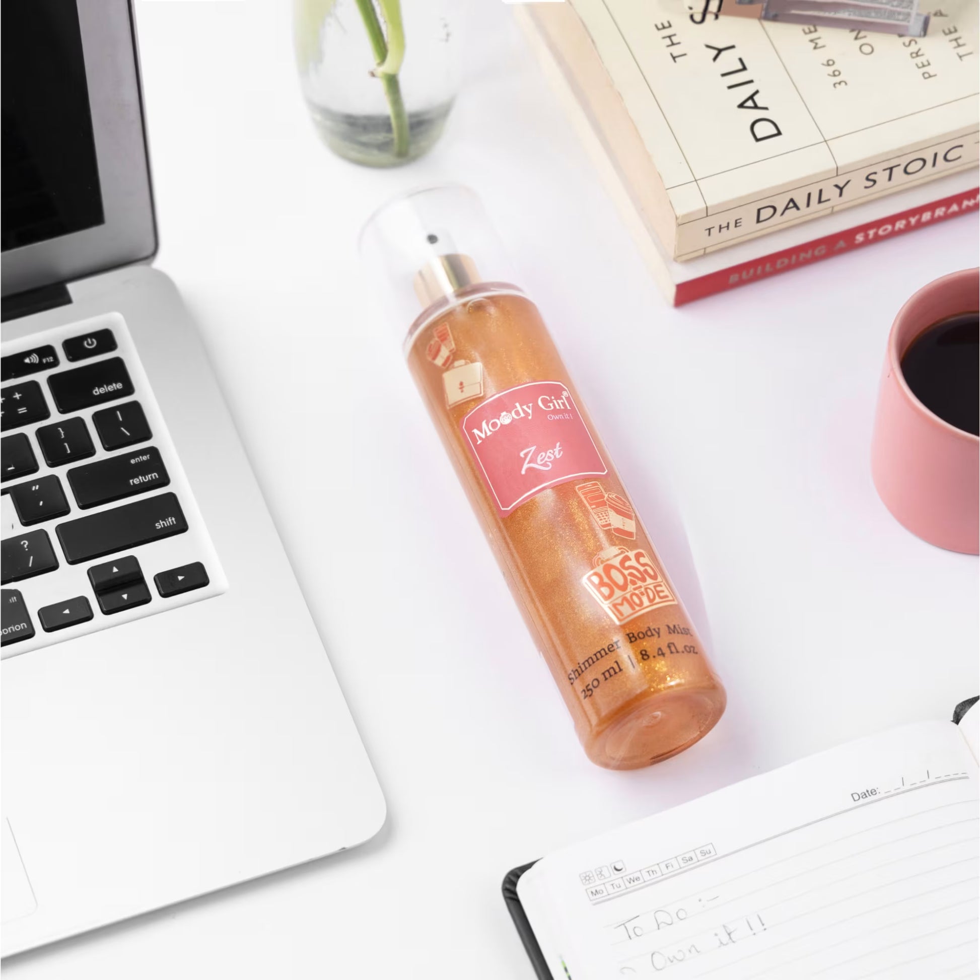 Bottle of 'Moody Girl Zest' body mist on a desk with a laptop, coffee cup, and books.