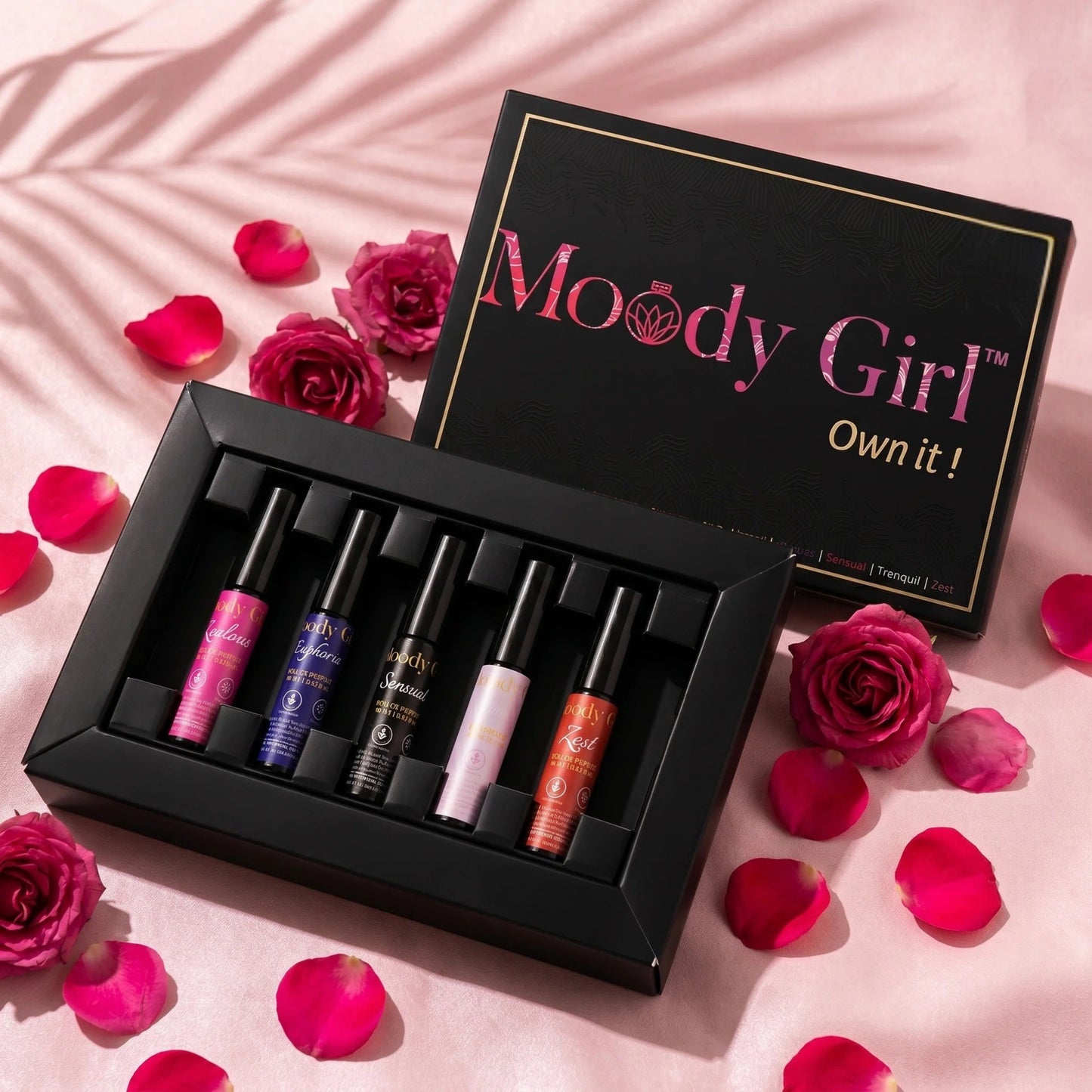 Set of lip glosses in a black box with 'Moody Girl' branding, surrounded by pink roses and petals on a pink background.