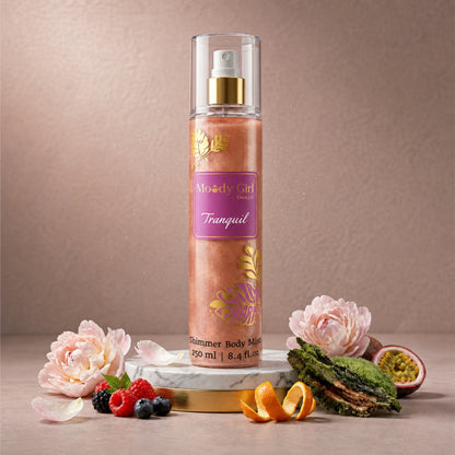 A bottle of Moody Girl Tranquil Shimmer Body Mist stands on a marble slab with decorative flowers, berries, citrus, and greenery around it against a soft beige background.